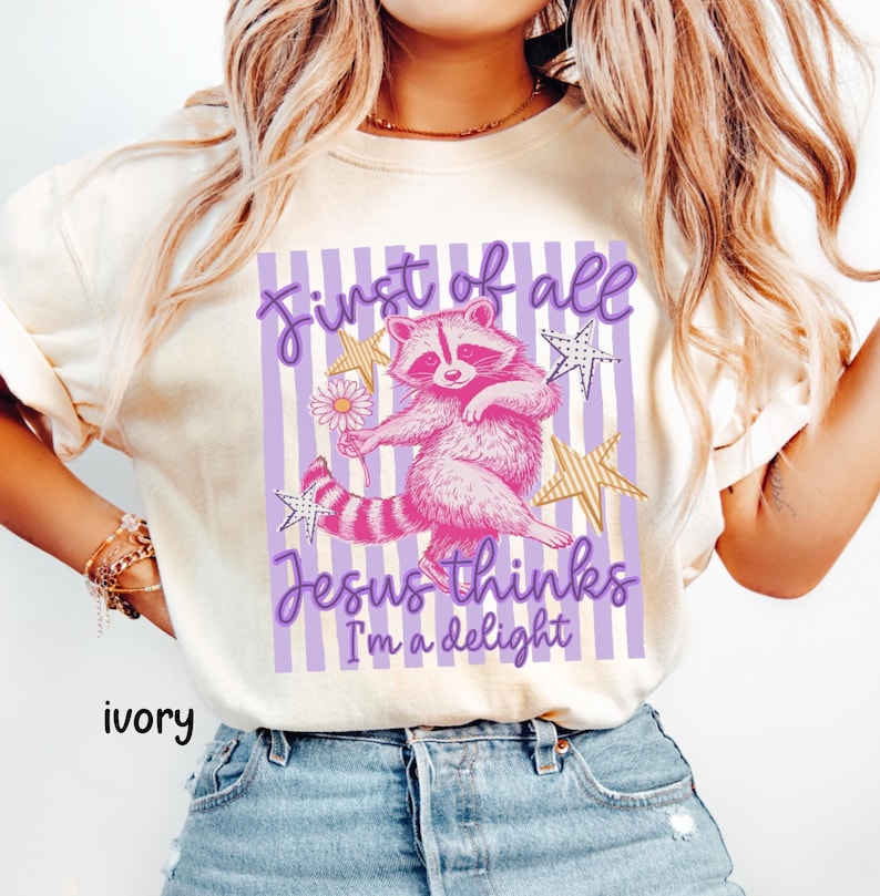 May include: Ivory-colored t-shirt featuring a pink raccoon illustration holding a flower, with text reading "First of all Jesus thinks I'm a delight." The design includes purple stripes and gold stars.