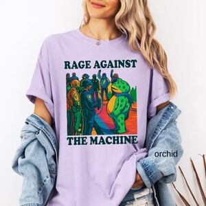 May include: Orchid-colored t-shirt with the text "RAGE AGAINST THE MACHINE" above a cartoon illustration. The illustration depicts a frog facing a group of police officers. The frog is wearing a scarf.