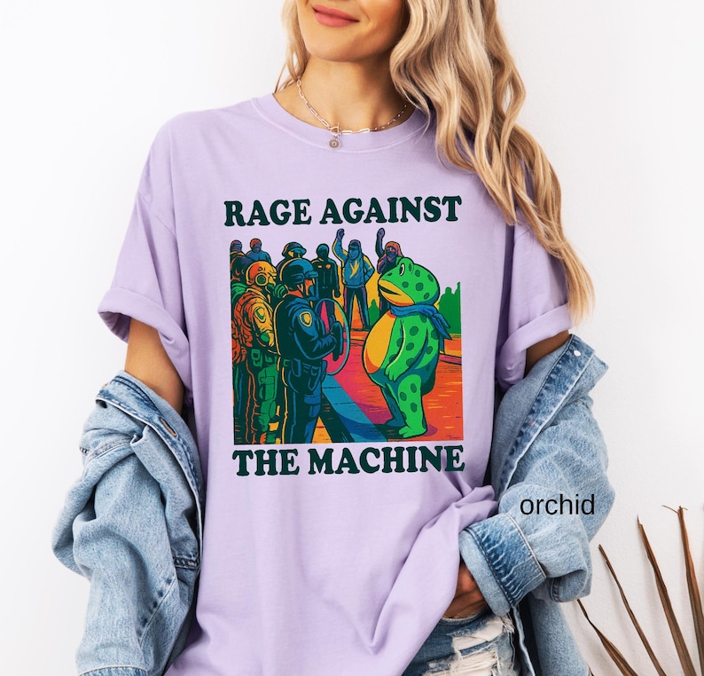 May include: Orchid-colored t-shirt with the text "RAGE AGAINST THE MACHINE" above a cartoon illustration. The illustration depicts a frog facing a group of police officers. The frog is wearing a scarf.