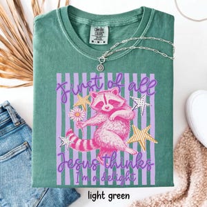 May include: Light green t-shirt with a graphic of a pink raccoon holding a flower, set against a purple striped background. Text reads "First of all Jesus thinks I'm a delight." The shirt has a crew neck and short sleeves.