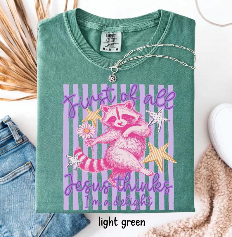 May include: Light green t-shirt with a graphic of a pink raccoon holding a flower, set against a purple striped background. Text reads "First of all Jesus thinks I'm a delight." The shirt has a crew neck and short sleeves.