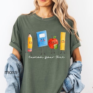 May include: A moss green t-shirt featuring a graphic of four anthropomorphic school supplies: a yellow pencil, a blue spiral notebook, a red apple with glasses holding a small green book, and a yellow ruler, with the text Custom Your Text.