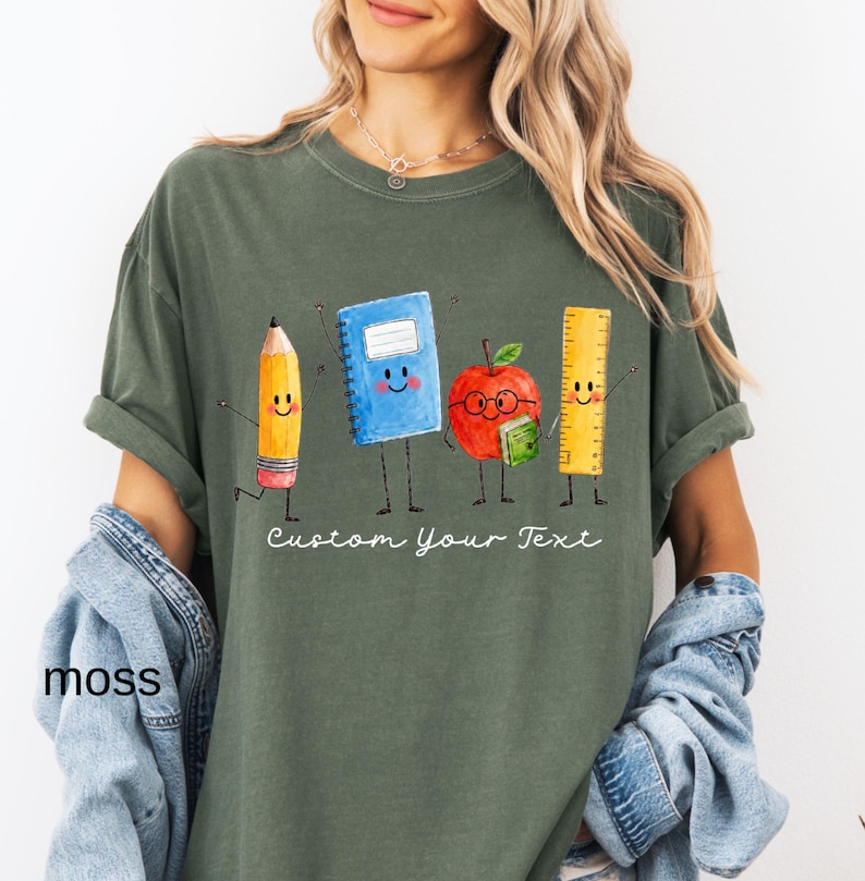 May include: A moss green t-shirt featuring a graphic of four anthropomorphic school supplies: a yellow pencil, a blue spiral notebook, a red apple with glasses holding a small green book, and a yellow ruler, with the text Custom Your Text.