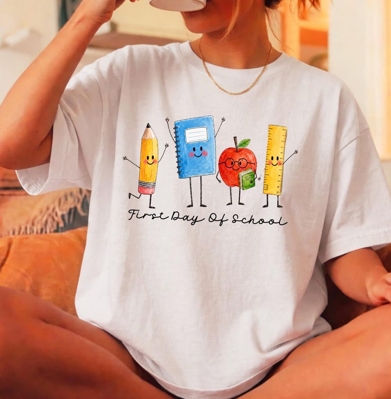 May include: A white t-shirt featuring a graphic of a yellow pencil, a blue notebook, a red apple with glasses, and a yellow ruler, all with arms and legs. Below the characters, the text First Day Of School is printed in a black cursive font.
