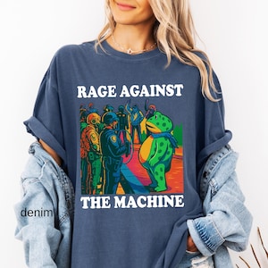 May include: Navy blue t-shirt with the text "RAGE AGAINST THE MACHINE" in white. The graphic features a green frog facing a group of police officers. The frog wears a blue scarf. The shirt is denim.