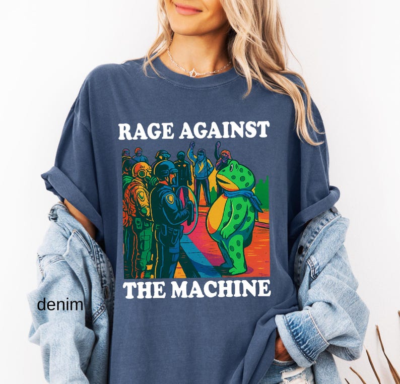 May include: Navy blue t-shirt with the text "RAGE AGAINST THE MACHINE" in white. The graphic features a green frog facing a group of police officers. The frog wears a blue scarf. The shirt is denim.