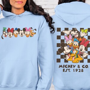 Puede incluir: Sudadera con capucha azul claro con caras de personajes de dibujos animados en el pecho. La parte trasera presenta un patr&oacute;n de cuadros marrones y blancos con personajes de dibujos animados y el texto "Mickey & Co. Est. 1928."