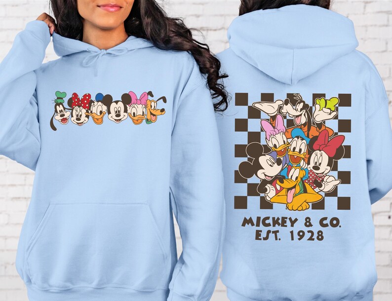 Puede incluir: Sudadera con capucha azul claro con caras de personajes de dibujos animados en el pecho. La parte trasera presenta un patr&oacute;n de cuadros marrones y blancos con personajes de dibujos animados y el texto "Mickey & Co. Est. 1928."