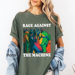 May include: Olive green t-shirt with the text "RAGE AGAINST THE MACHINE" in white. The graphic depicts a frog in a scarf facing a group of police officers. The shirt is a relaxed fit.