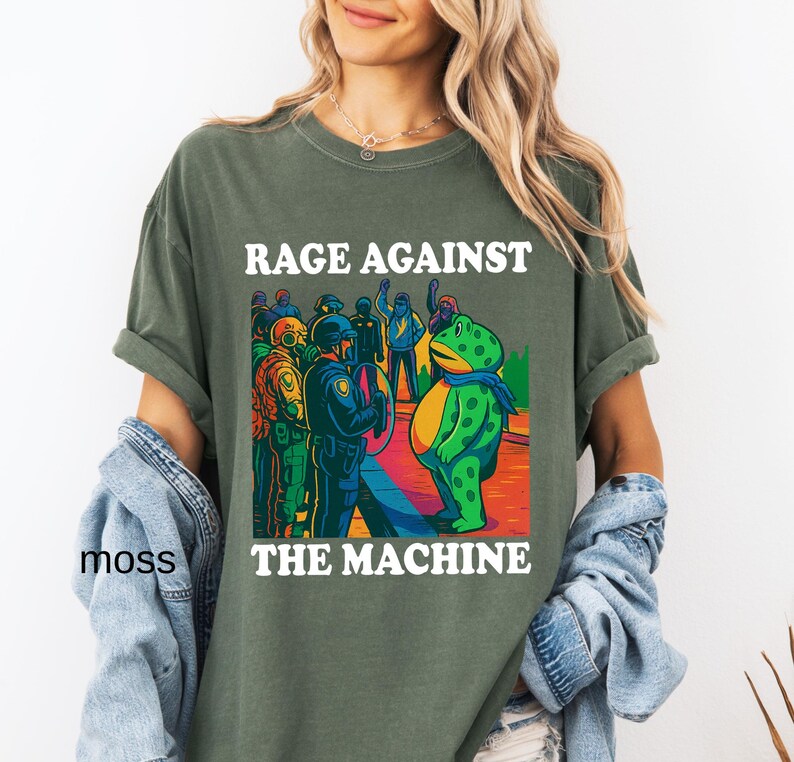 May include: Olive green t-shirt with the text "RAGE AGAINST THE MACHINE" in white. The graphic depicts a frog in a scarf facing a group of police officers. The shirt is a relaxed fit.
