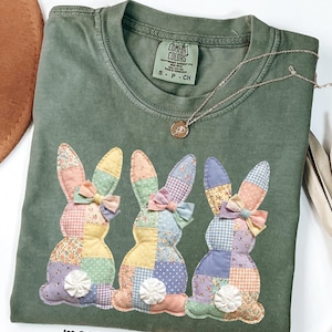 May include: Moss green t-shirt with three patchwork bunny rabbit appliqués. Each bunny has a different patterned ear and bow. The shirt has a crew neck and short sleeves. A silver necklace with a small pendant is draped across the shirt.