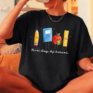 May include: A black t-shirt featuring a graphic of a yellow pencil, a blue notebook, a red apple with glasses, and a yellow ruler, all with smiling faces and stick arms. The text First Day Of School is printed in white below the characters.