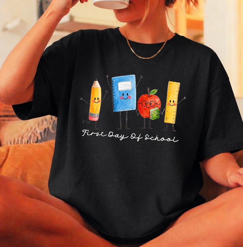 May include: A black t-shirt featuring a graphic of a yellow pencil, a blue notebook, a red apple with glasses, and a yellow ruler, all with smiling faces and stick arms. The text First Day Of School is printed in white below the characters.