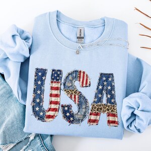 May include: A light blue sweatshirt with the word "USA" appliqued on the front. The letters are made of denim, leopard print, and the American flag. A silver necklace with a sun pendant is on the sweatshirt.