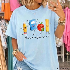 May include: A light blue t-shirt featuring a graphic of a yellow pencil, a blue notebook, a red apple with glasses, and a yellow ruler, all with smiling faces and arms. The word "Kindergarten" is printed in black cursive script below the characters.