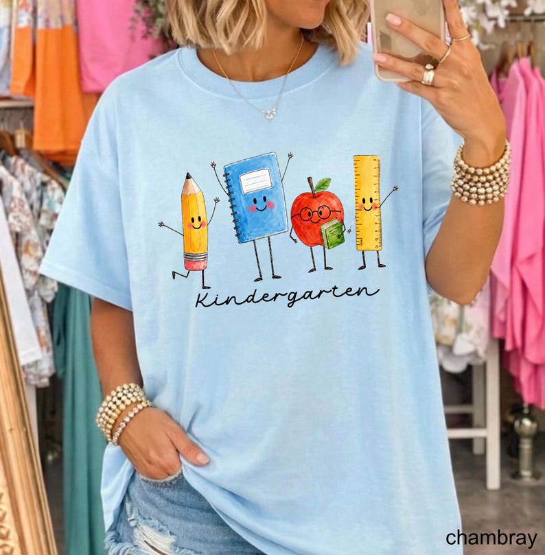 May include: A light blue t-shirt featuring a graphic of a yellow pencil, a blue notebook, a red apple with glasses, and a yellow ruler, all with smiling faces and arms. The word "Kindergarten" is printed in black cursive script below the characters.