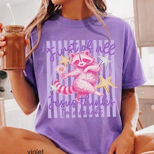 May include: Violet t-shirt featuring a whimsical raccoon illustration holding a flower and surrounded by stars. The text reads "First of all Jesus thinks I'm a delight". The shirt is a comfortable, casual wear item.