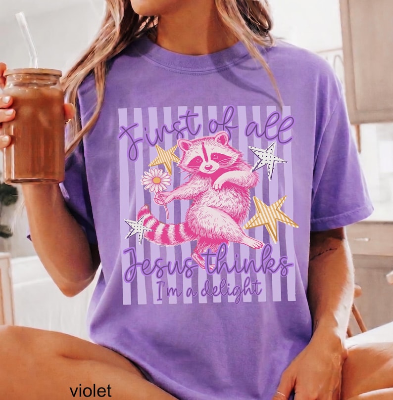 May include: Violet t-shirt featuring a whimsical raccoon illustration holding a flower and surrounded by stars. The text reads "First of all Jesus thinks I'm a delight". The shirt is a comfortable, casual wear item.