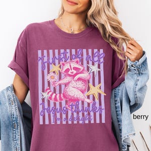 May include: Berry-colored t-shirt featuring a pink raccoon holding a daisy, with the text "First of all Jesus thinks I'm a delight." The design includes gold star accents and purple vertical stripes.