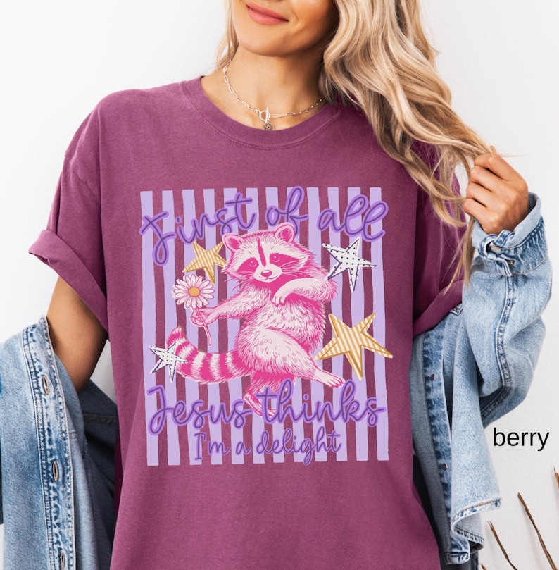 May include: Berry-colored t-shirt featuring a pink raccoon holding a daisy, with the text "First of all Jesus thinks I'm a delight." The design includes gold star accents and purple vertical stripes.