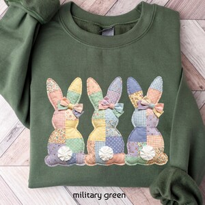 May include: Military green sweatshirt featuring three appliqued bunny rabbits. Each bunny has patchwork-style bodies in pastel colors, with a bow and a white flower. The text "military green" is at the bottom of the image.