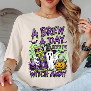 May include: A cream-colored t-shirt featuring a graphic print with the text A BREW A DAY KEEPS THE WITCH AWAY. The design includes a ghost wearing a witch hat, a purple energy drink can labeled WITCH'S BREW, a pumpkin, and small purple bats.