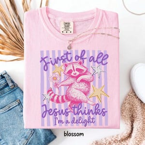 May include: Pink Comfort Colors t-shirt with a raccoon graphic holding a flower, surrounded by stars and the text "First of all Jesus thinks I'm a delight". The word "blossom" is at the bottom.