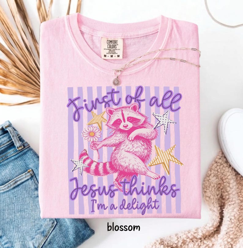 May include: Pink Comfort Colors t-shirt with a raccoon graphic holding a flower, surrounded by stars and the text "First of all Jesus thinks I'm a delight". The word "blossom" is at the bottom.