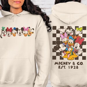 Puede incluir: Sudadera con capucha color crema con un gr&aacute;fico colorido de personajes de dibujos animados en la parte delantera y trasera. La parte delantera muestra caras de personajes, mientras que la parte trasera muestra un grupo de personajes sobre un fondo a cuadros. El texto "Mickey & Co. EST. 1928" est&aacute; impreso debajo.