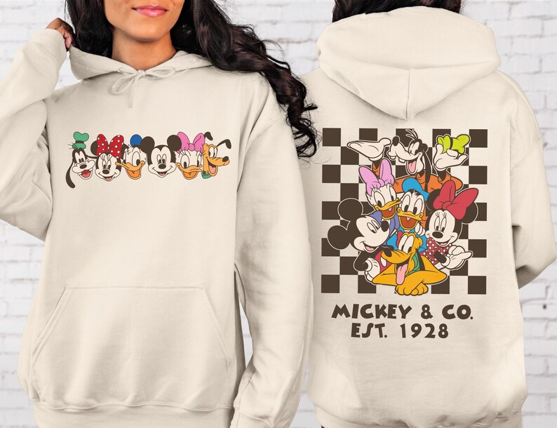 Puede incluir: Sudadera con capucha color crema con un gr&aacute;fico colorido de personajes de dibujos animados en la parte delantera y trasera. La parte delantera muestra caras de personajes, mientras que la parte trasera muestra un grupo de personajes sobre un fondo a cuadros. El texto "Mickey & Co. EST. 1928" est&aacute; impreso debajo.