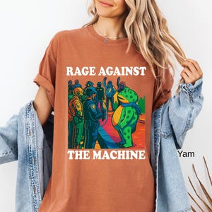 May include: Rust-colored t-shirt with the text "RAGE AGAINST THE MACHINE" in white. The graphic features a green frog with a blue scarf facing a group of police officers. The frog is standing on a colorful, rainbow-like surface.