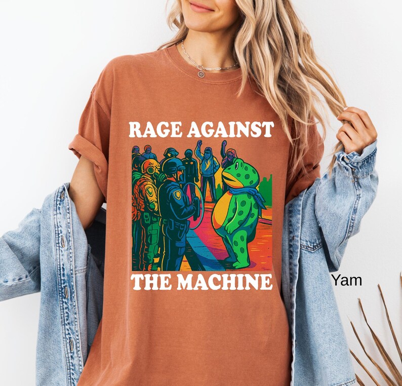 May include: Rust-colored t-shirt with the text "RAGE AGAINST THE MACHINE" in white. The graphic features a green frog with a blue scarf facing a group of police officers. The frog is standing on a colorful, rainbow-like surface.