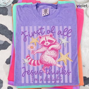 May include: A stack of colorful t-shirts, with a violet shirt on top. The violet shirt features a raccoon graphic holding a flower, with text that reads "First of all Jesus thinks I'm a delight."
