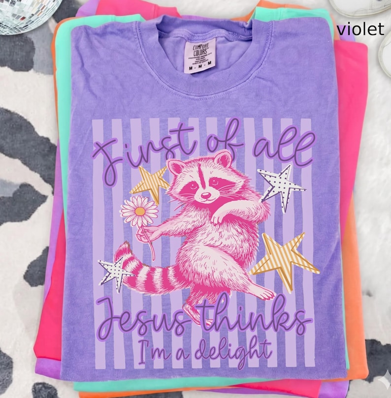May include: A stack of colorful t-shirts, with a violet shirt on top. The violet shirt features a raccoon graphic holding a flower, with text that reads "First of all Jesus thinks I'm a delight."