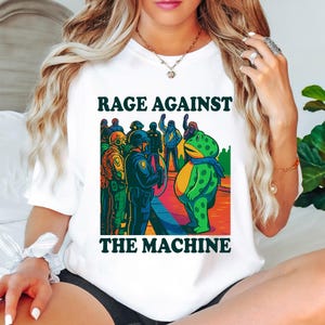 May include: White t-shirt with the text "RAGE AGAINST THE MACHINE" in a bold, dark green font. The graphic features a cartoon frog facing a line of police officers. The frog is green with yellow spots and a blue scarf.