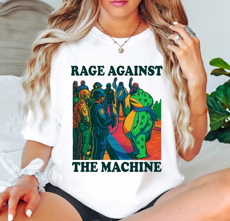 May include: White t-shirt with the text "RAGE AGAINST THE MACHINE" in a bold, dark green font. The graphic features a cartoon frog facing a line of police officers. The frog is green with yellow spots and a blue scarf.