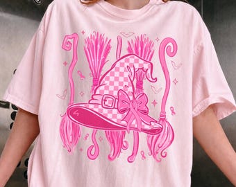 Può includere: Una t-shirt rosa chiaro con una stampa grafica di un cappello da strega a scacchi decorato con un fiocco e un nastro. Il design include due scope, piccoli pipistrelli e accenti a forma di stella in varie tonalità di rosa.