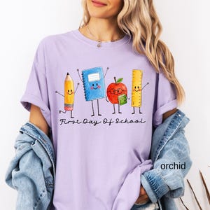 May include: A light purple t-shirt featuring a graphic of a cartoon pencil, blue notebook, red apple with glasses, and yellow ruler. Below the characters, the text reads First Day Of School in black cursive script. The shirt has short sleeves.