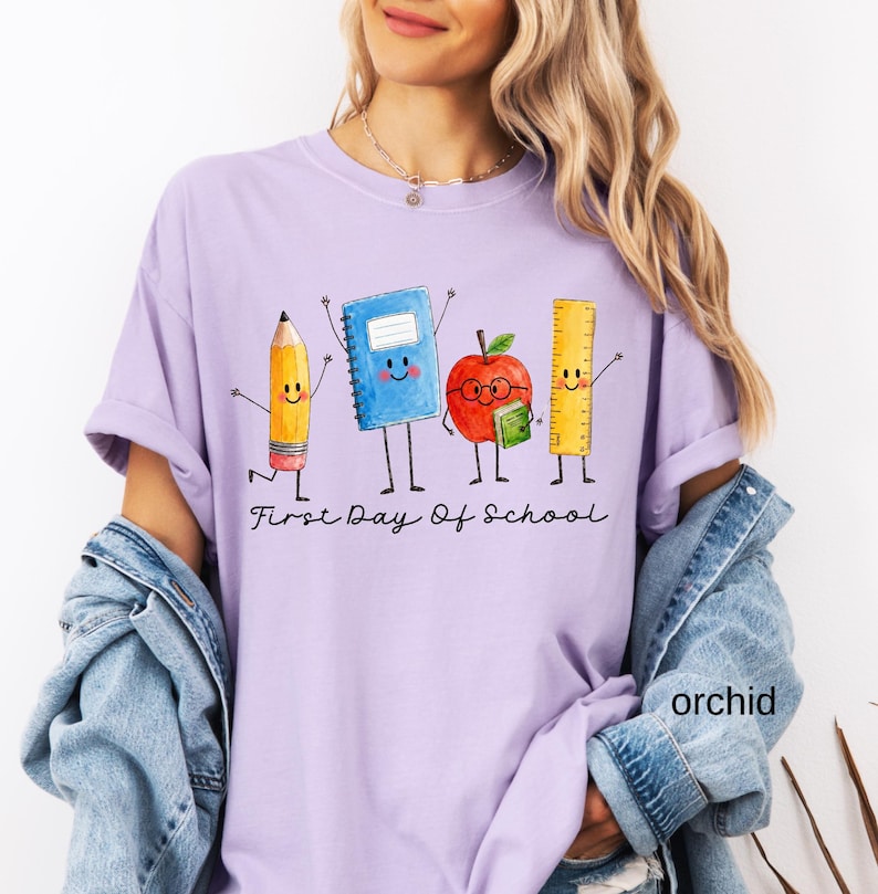 May include: A light purple t-shirt featuring a graphic of a cartoon pencil, blue notebook, red apple with glasses, and yellow ruler. Below the characters, the text reads First Day Of School in black cursive script. The shirt has short sleeves.