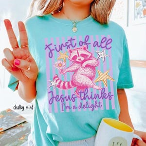 May include: A light turquoise t-shirt with a graphic of a pink raccoon holding a daisy, surrounded by stars and vertical stripes. Text reads "First of all Jesus thinks I'm a delight."