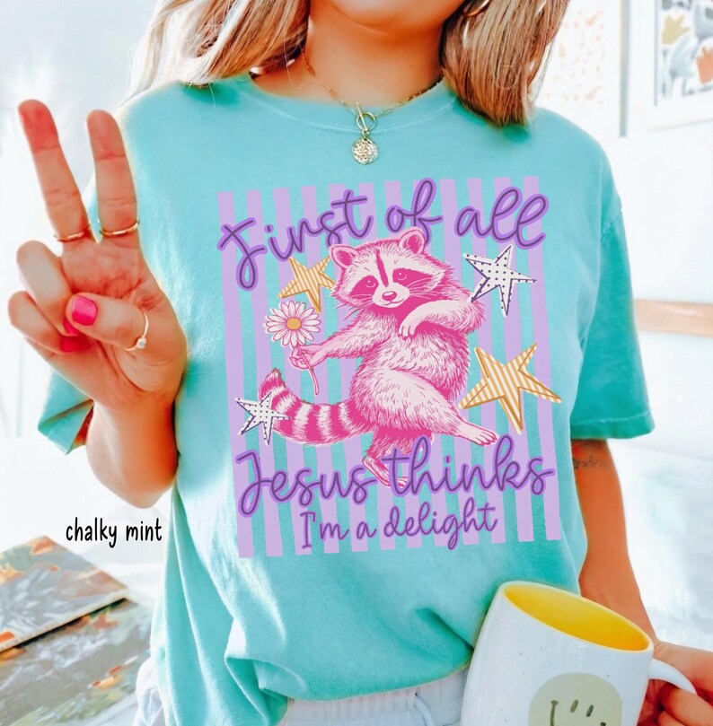 May include: A light turquoise t-shirt with a graphic of a pink raccoon holding a daisy, surrounded by stars and vertical stripes. Text reads "First of all Jesus thinks I'm a delight."