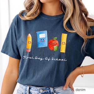May include: A dark blue t-shirt featuring a graphic of a smiling pencil, a blue notebook, a red apple with glasses, and a yellow ruler. Below the characters, the white text reads First Day Of School in a cursive script font style.