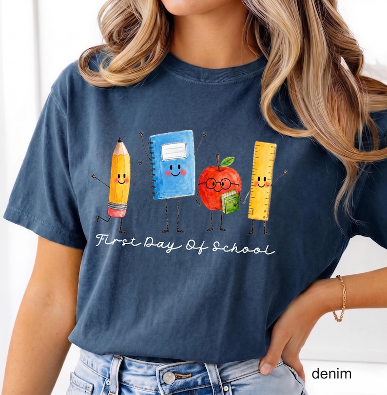 May include: A dark blue t-shirt featuring a graphic of a smiling pencil, a blue notebook, a red apple with glasses, and a yellow ruler. Below the characters, the white text reads First Day Of School in a cursive script font style.