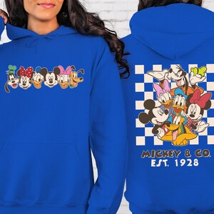 Puede incluir: Sudadera con capucha azul real con un dise&ntilde;o de personajes de Disney. La parte delantera muestra caras de personajes, mientras que la parte trasera muestra un grupo de personajes sobre un fondo a cuadros con "Mickey & Co. Est. 1928".