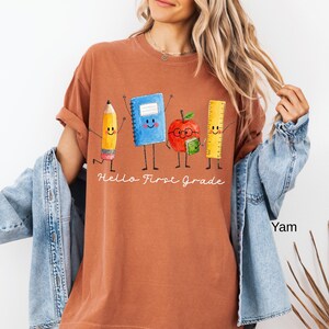 May include: A rust-colored t-shirt featuring a graphic of a smiling pencil, blue notebook, red apple with glasses, and yellow ruler. Below the characters, the text reads "Hello First Grade" in white cursive script. The shirt has short sleeves.