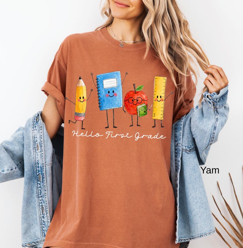 May include: A rust-colored t-shirt featuring a graphic of a smiling pencil, blue notebook, red apple with glasses, and yellow ruler. Below the characters, the text reads "Hello First Grade" in white cursive script. The shirt has short sleeves.