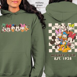 Puede incluir: Sudadera con capucha verde militar con un gr&aacute;fico frontal de caras de personajes de dibujos animados y un gr&aacute;fico trasero de los mismos personajes sobre un fondo a cuadros. El gr&aacute;fico trasero incluye el texto "MICKEY & CO. EST. 1928". La sudadera tiene una capucha con cord&oacute;n y un bolsillo delantero.