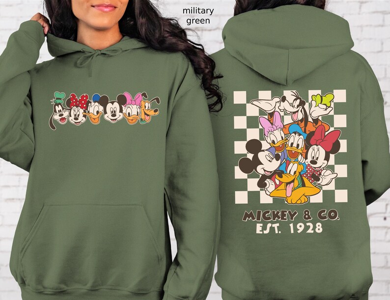 Puede incluir: Sudadera con capucha verde militar con un gr&aacute;fico frontal de caras de personajes de dibujos animados y un gr&aacute;fico trasero de los mismos personajes sobre un fondo a cuadros. El gr&aacute;fico trasero incluye el texto "MICKEY & CO. EST. 1928". La sudadera tiene una capucha con cord&oacute;n y un bolsillo delantero.