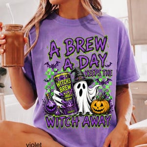 May include: A purple t-shirt featuring a graphic print with the text A BREW A DAY KEEPS THE WITCH AWAY. The design includes a ghost wearing a witch hat, a can labeled WITCH'S BREW, a pumpkin, small purple bats, and a green checkered background.