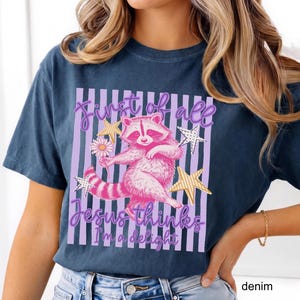 May include: Denim blue t-shirt featuring a pink raccoon holding a daisy, with the text "First of all Jesus thinks I'm a delight" in a purple and white striped design. The design includes gold star accents.